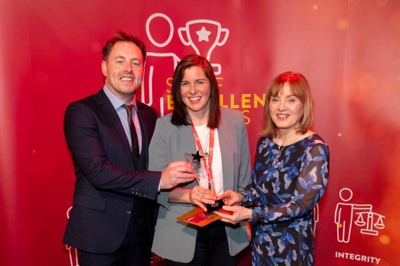 A man and three women standing in front of a large red Staff Excellence Awards banner. Lady in the middle is hold a glass trophy award