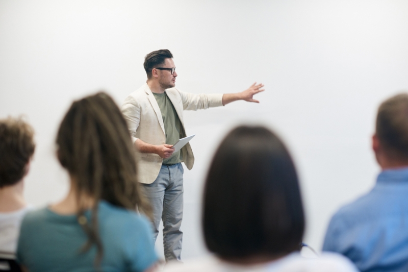 young man holding notepad and gesturing to his left in a lecture or presentation setting