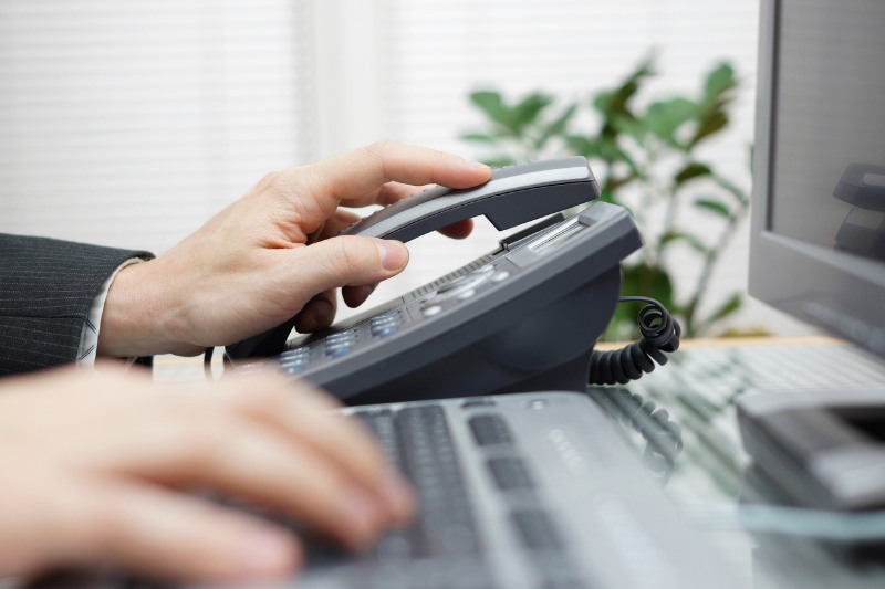 businessman working at office desk placing one hand on a phone and the other on a computer keyboard